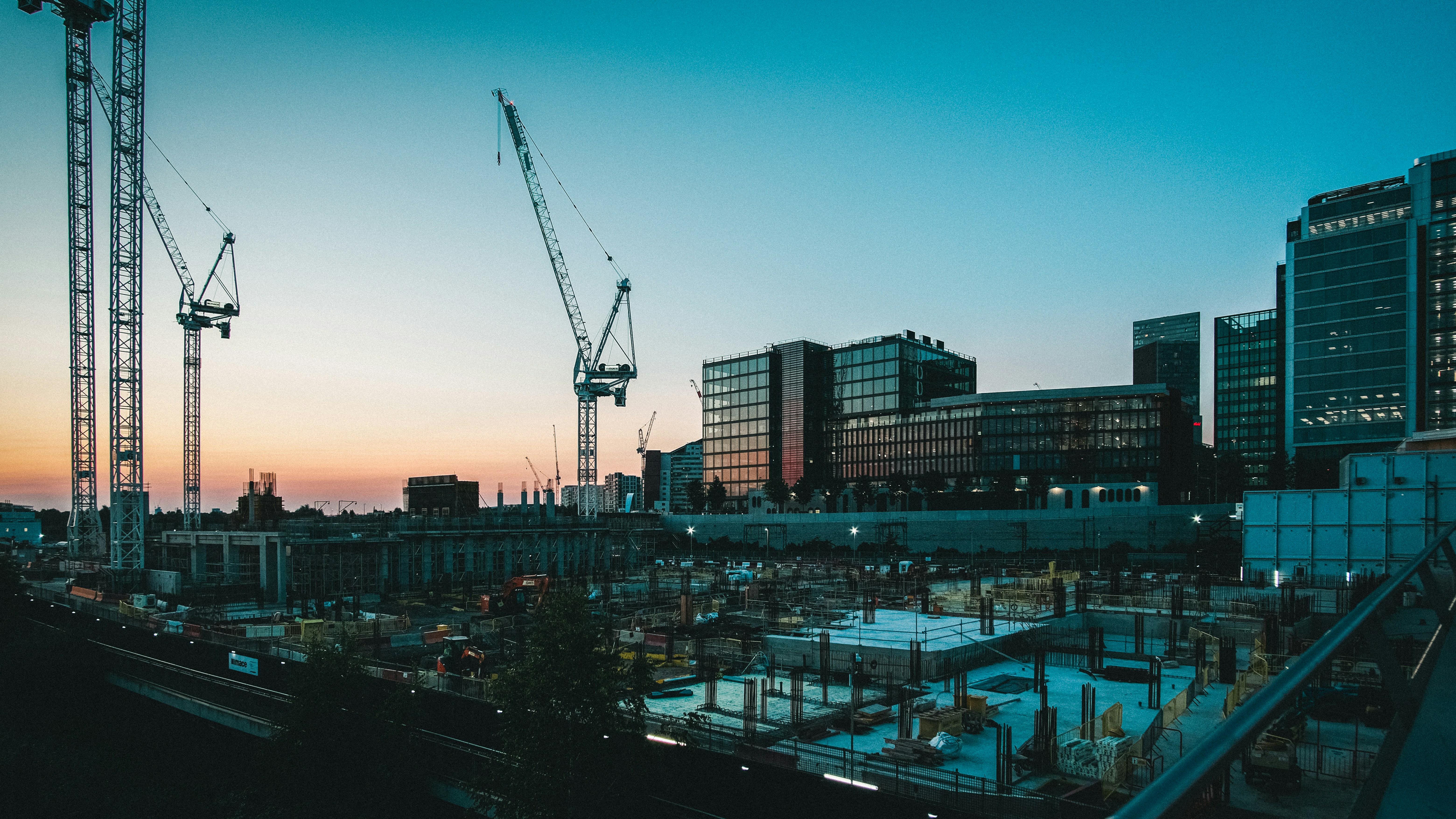 Urban construction site at dusk – concrete foundation work with steel rebar, tower cranes, and illuminated glass office towers against a twilight sky.