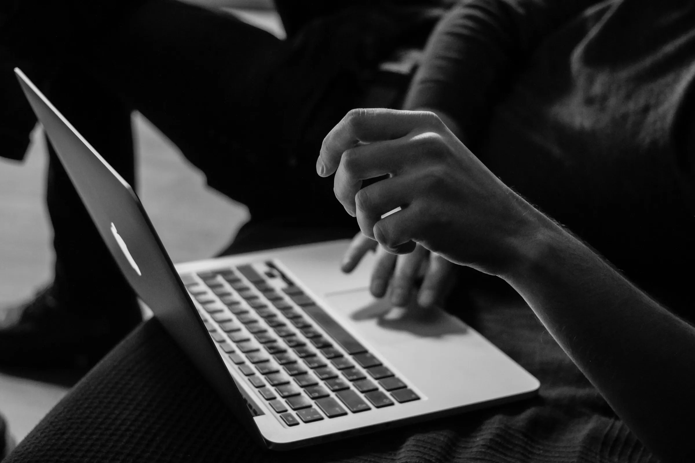 Grayscale photograph of a person working at a laptop on a wooden desk, hands resting on the keyboard.
