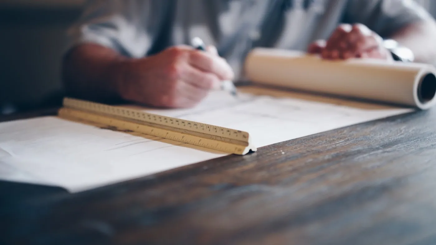 Close-up of hands drafting on architectural drawings with a scale ruler on a wooden desk – hands-on planning and coordinated execution. Stand-in (Unsplash, Daniel McCullough; see public/assets/platform/LICENSES.md).