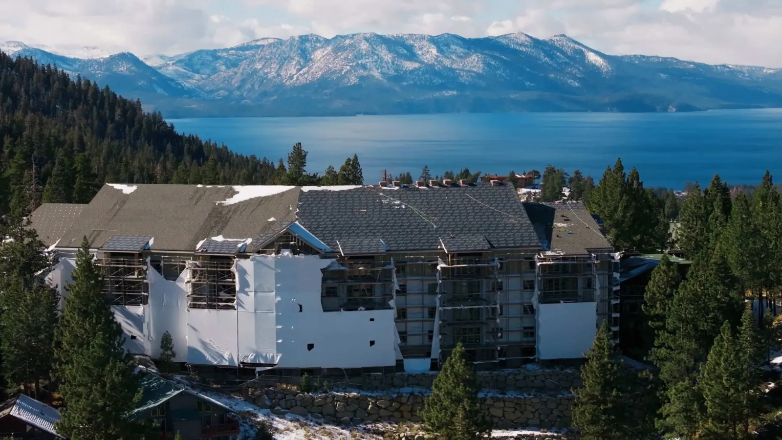 The Peak, South Lake Tahoe – the multi-story luxury condominium under construction, wrapped in scaffolding and weather sheeting above Lake Tahoe with the snow-capped Sierra Nevada beyond. New Course Enterprises project imagery.
