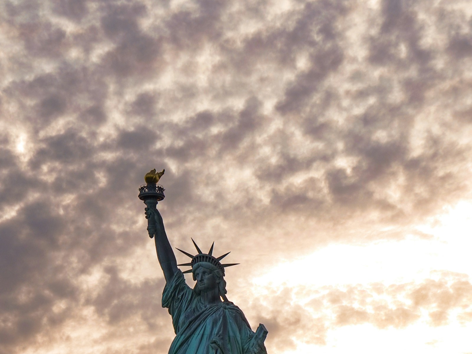 The Statue of Liberty silhouetted against a cloudy sky in New York Harbor – Bobby's HOME-P2 thematic spec for EB-5 ('reference to immigration'). Statue of Liberty is a US federal public monument (not a trademark); no commercial brand marks in frame. Stand-in (Unsplash, Koushalya Karthikeyan; see public/assets/platform/LICENSES.md).