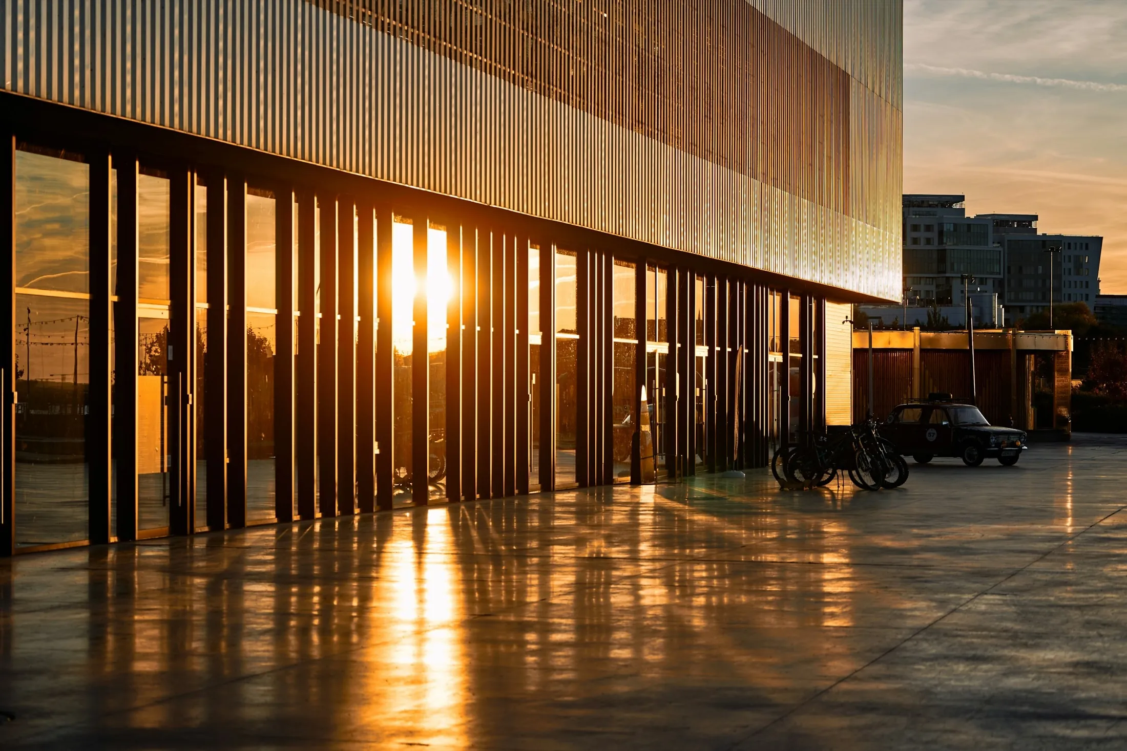 Contemporary building colonnade at golden hour – low sun flaring through vertical facade fins across a mirror-smooth reflecting plaza. Premium architectural stand-in for the /platform overview hero (Unsplash, Maxim Tolchinskiy; see public/assets/platform/LICENSES.md). Owned NCE photography is the DESIGN.md §17 swap target.