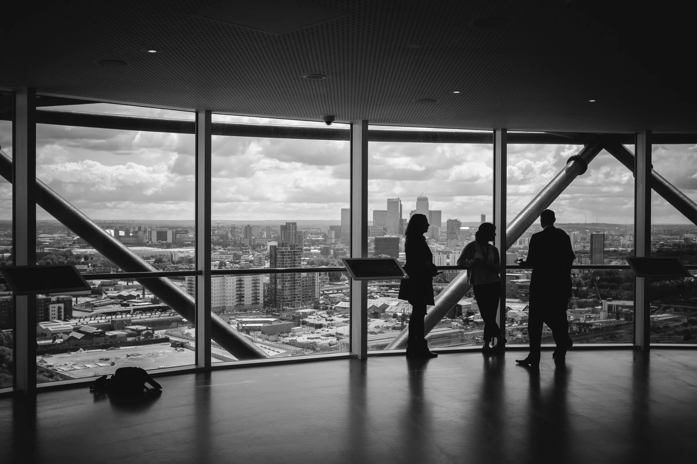 Three colleagues in silhouette talking by the floor-to-ceiling windows of a high-rise office, a monochrome city skyline stretching to the horizon beyond.